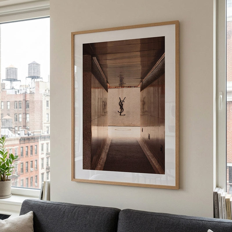 A framed art print featuring a sepia-toned photograph of a black YSL logo sculpture in a hallway, displayed on a beige wall above a dark gray sofa in a modern living room setting.