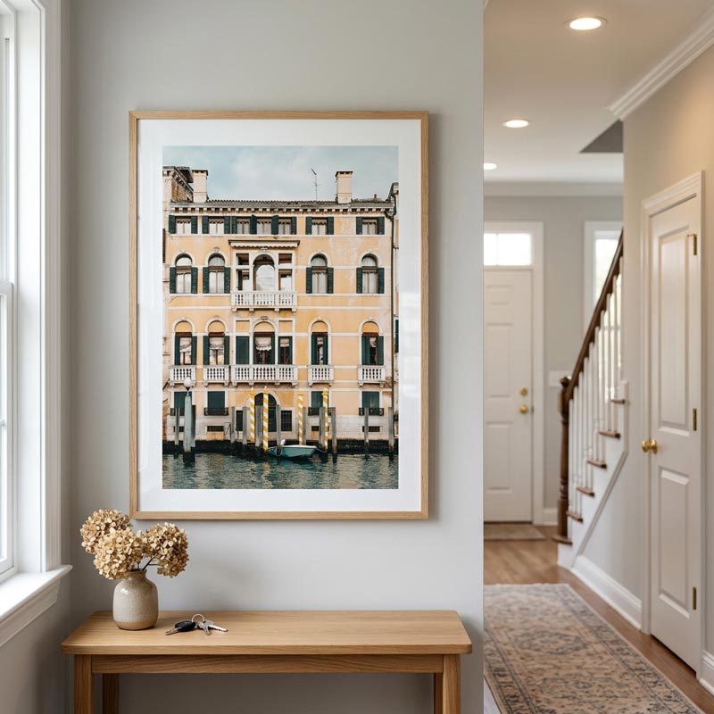 A framed art print of a Venetian canal building hangs on a light gray wall above a wooden console table in a bright, modern hallway.