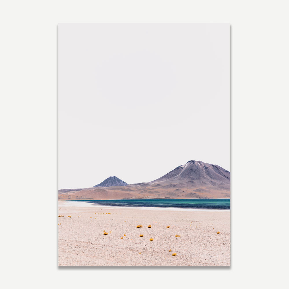 A landscape photograph featuring a body of water, likely a lagoon, with mountains in the background under a clear sky. The foreground includes a desert-like area with scattered yellow objects.